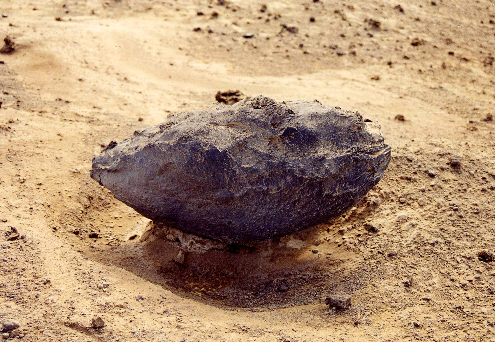 Zdjęcie przedstawia bombę wulkaniczną. Ma wydłużony kształt o nieregularnej i ciemnej powierzchni. Leży na piasku.
