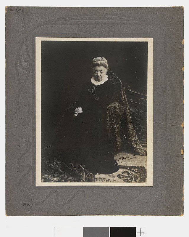 Zdjęcie przedstawia portret siedzącej na krześle starszej kobiety. Postać ukazuje trzy czwarte twarzy. Kobieta obraca głowę w swoją lewą stronę. Jej twarz jest owalna. Okalają ją zaczesane ku górze i upięte na czubku długie włosy. Kobieta ma zmarszczone, niskie czoło. Brwi są wyraziste. Jej powieki lekko opadają. Patrzy na nas. Jej nos jest prosty, mocno zarysowany. Ma wydatne usta. Pod szyją ma koronkową, małą kryzę, która wystaje z dekoltu sukni. Kobieta składa dłonie na kolanach.