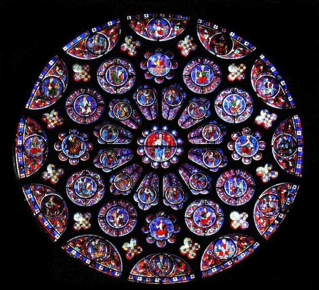 Zdjęcie przedstawia rozetę z witrażami w Katedrze Chartres. W centralnej części rozety znajduje się Jezus Chrystus. Rozeta jest symetryczna i wielokolorowa. Poszczególne elementy rozety są podzielone na pola w kształcie okręgów, półokręgów oraz form o kształcie wydłużonego owalu zwężającego się z jednej strony. Pomiędzy tymi polami znajduje się czarne tło.