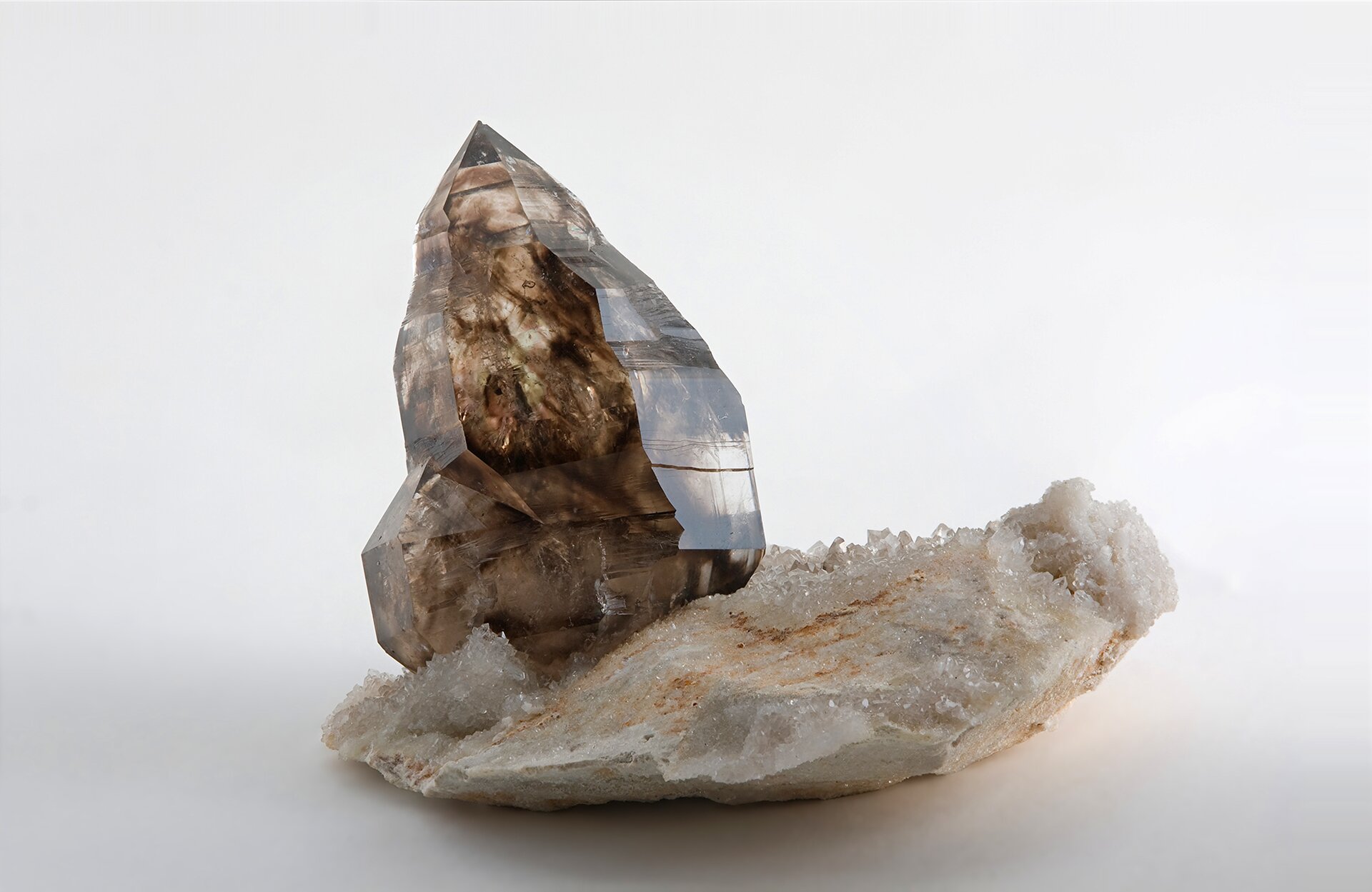 Zdjęcie przedstawia brązowy kryształ kwarcu dymnego w postaci pojedynczego słupka na białej podstawie mineralnej pokrytej drobnymi białymi kryształkami. Tło białe.