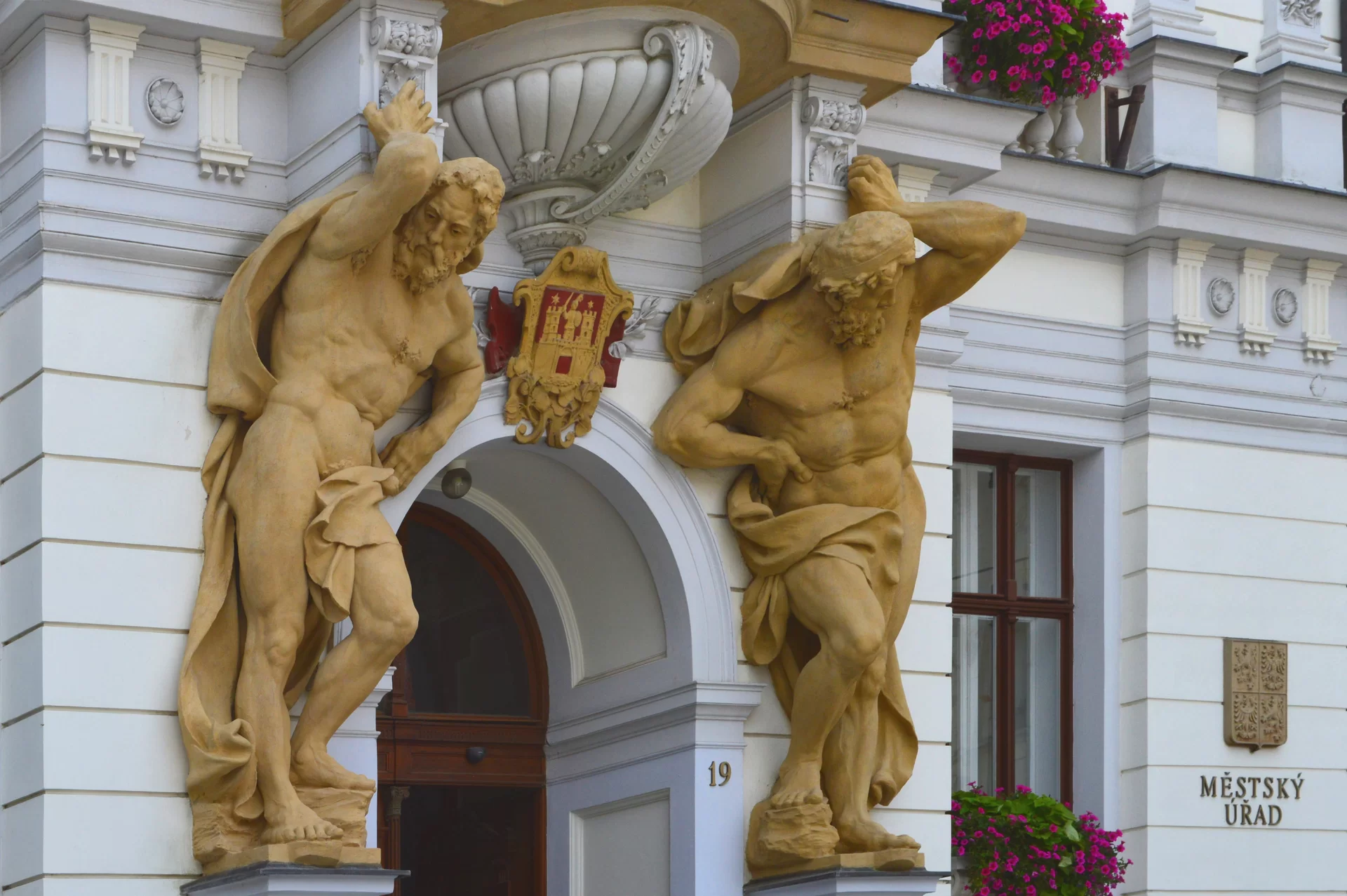Zdjęcie przedstawia fragment budynku. W przednią ścianę budynku wbudowano portal o łukowym sklepieniu, który prowadzi do drewnianych drzwi. Po obu stronach portalu umieszczono żółte rzeźby dwóch stojących mężczyzn. Są oni nadzy, przewiązani w pasie chustą, mają zarost na twarzy. Podtrzymują na barkach element umieszczony nad nimi.