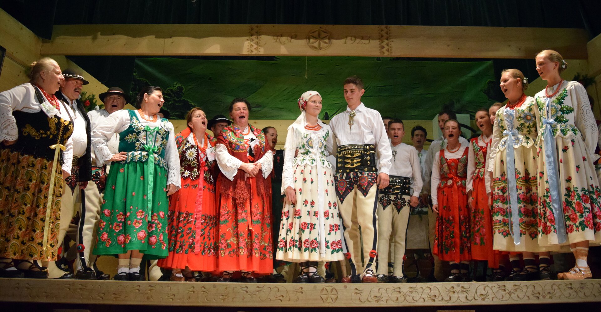 Grupa górali podhalańskich. Pośrodku młoda dziewczyna i chłopak. Około dwudziestoletni, noszący tradycyjne stroje. Noszą charakterystyczne kierpce, kobieta ma długą, białą suknię z wyszywanymi na niej różami, białą bluzkę i chustę na głowie. Mężczyzna białą koszulę, spodnie z haftowanymi ozdobami u góry oraz szeroki pas. Wokół nich grupa górali w różnym wieku i różnej płci, również ubranych w tradycyjne stroje. Wszyscy razem śpiewają.