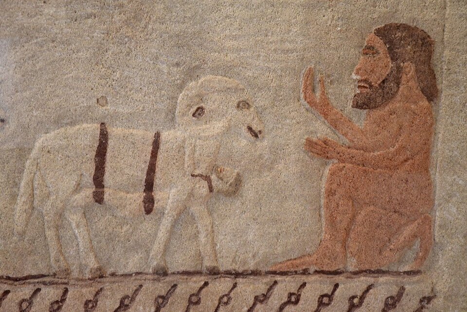 Kolorowa fotografia przedstawia płaskorzeźbę na kamieniu. Po prawej stronie przyklęknął na prawym kolanie ogromny mężczyzna z brodą, to Polifem. Uniósł prawą rękę do góry. Przed nim stoi rogaty baran, pod jego brzuchem znajduje się przywiązany dwoma sznurami człowiek.