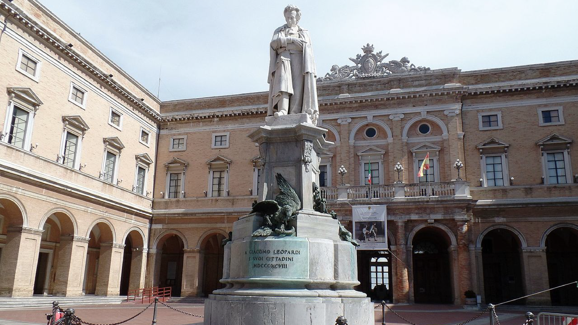 Zdjęcie przedstawia budynek ratuszu w miejscowości Recanati. Przed budynkiem ratusza, na pierwszym planie, widoczny jest pomnik, na którym znajduje się męska postać.