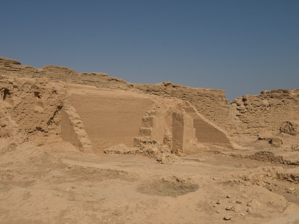 Ruiny starożytnej budowli z suszonej cegły na pustynnym terenie: niskie, częściowo zachowane ściany z widocznymi wnękami i schodkowym fragmentem muru. Jasnobrązowa ziemia i kamienie otaczają obiekt, a nad nim rozciąga się bezchmurne, błękitne niebo.
