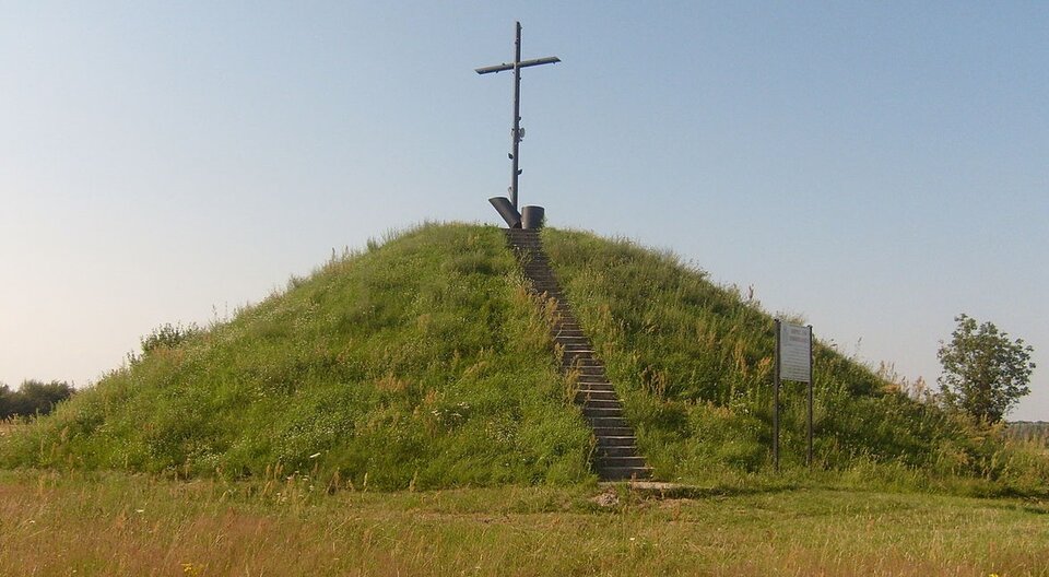 Zdjęcie przedstawia kopiec z krzyżem na szczycie. Na szczyt prowadzą betonowe schody.  