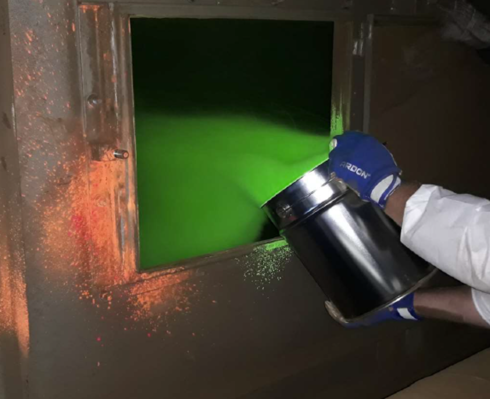 Zdjęcie prezentuje działania konserwacyjne polegające na napylaniu proszkiem fluorescencyjnym filtra workowego pieca hutniczego. Na obrazku znajduje się wiadro z proszkiem, które trzyma osoba ubrana w rękawiczki. Proszek zostaje wprowadzany do pieca hutniczego.