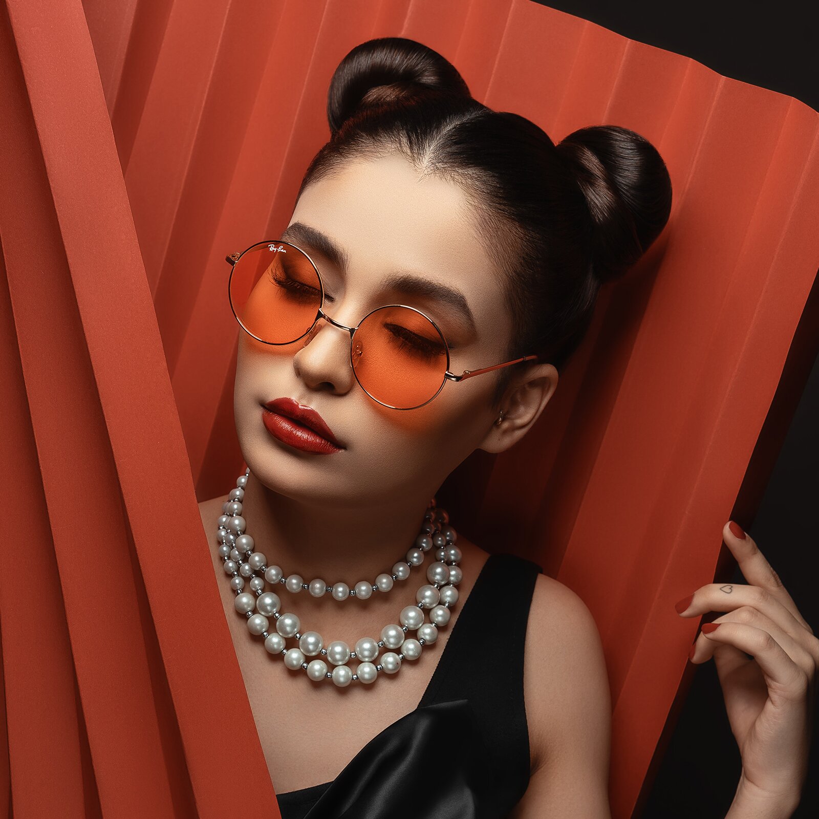 Zdjęcie przedstawia młodą kobietę. Ma brązowe włosy upięte w dwa koki. Ma długie rzęsy. Jej usta i paznokcie są pomalowane na czerwono. Nosi okulary przeciwsłoneczne. Na szyi ma korale.