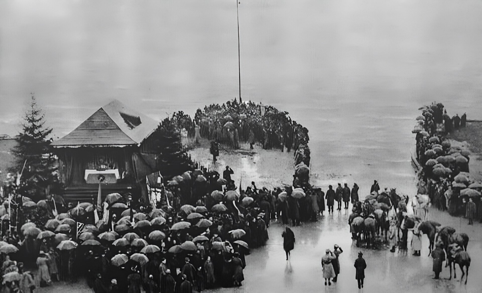Zdjęcie przedstawia tłum ludzi na przystani nadbrzeżnej w porcie puckim, w płaszczach i pod parasolami. Pierwsza część tłumu stoi w głębi kadru - na cyplu mola, na którego końcu znajduje się maszt. Na pierwszym planie, po lewej stronie kadru druga część tłumu, stłoczona, stoi pod budynkiem o czterospadowym dachu. Na drugim planie, po prawej stronie kadru, trzecia część tłumu spogląda na scenę w centrum fotografii z mola. Pojedyncze osoby widoczne są u dołu zdjęcia, na pierwszym planie. W tle widać wody Zatoki Puckiej. 