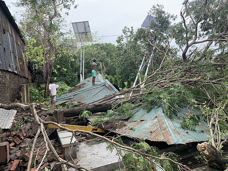 Zdjęcie przedstawiające zniszczony teren przez cyklon Amfan. Po lewej stronie ściana budynku, w centralnej części na ziemi leży zerwany blaszany dach, powalone drzewa, fragmenty zniszczonego budynku. Po powierzchni zerwanego dachu chodzą ludzie. Krajobraz tropikalny.