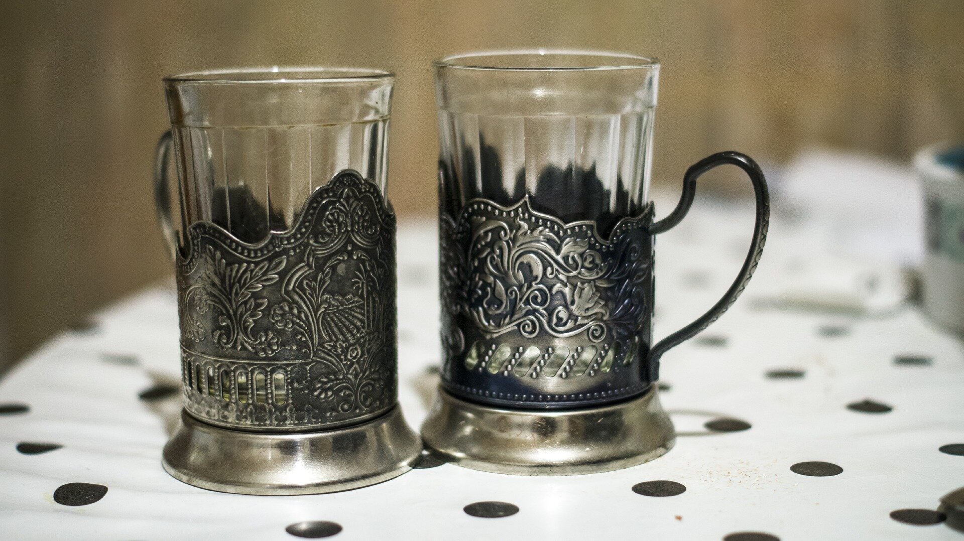 Na zdjęciu widoczne są dwie szklanki wykonane z cienkiego, przezroczystego szkła. Szklanki umieszczone są w metalowych koszyczkach z uszkami. Obydwa naczynia stoją na białym obrusie w czarne kropki.