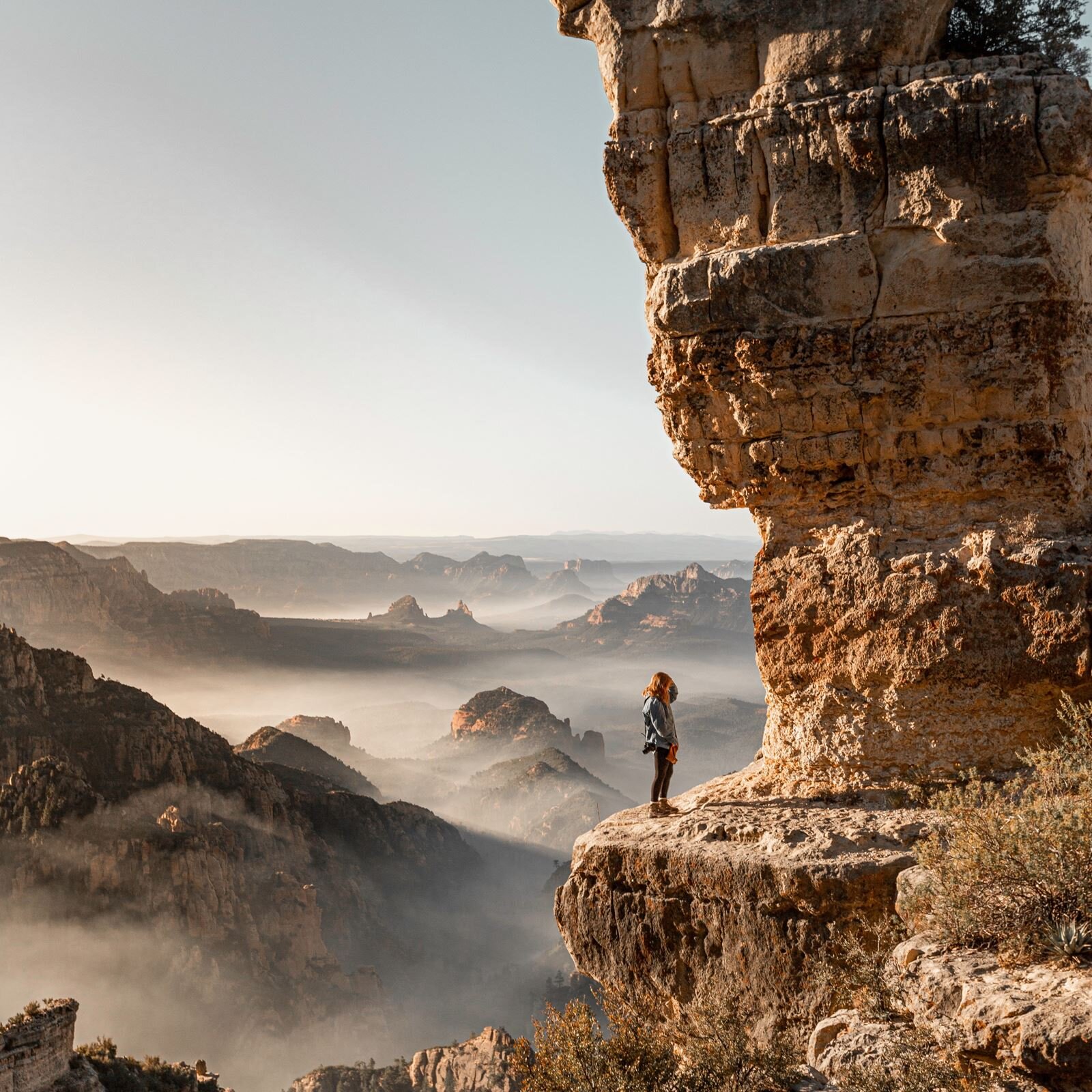 Zdjęcie przedstawia kobietę stojącą na półce skalnej. Kobieta stoi zwrócona w prawo, twarzą do wysokiej skały znajdującej się nad półką skalną. Kobieta ma długie, rude włosy, jest ubrana w niebieską kurtkę, czarne leginsy i górskie buty, a na prawym ramieniu ma przewieszony aparat fotograficzny. W prawej ręce trzyma czapkę, a lewą ręką dotyka głowy. W tle widać skaliste wzgórza, których dolne części są zasnute mgłą.
