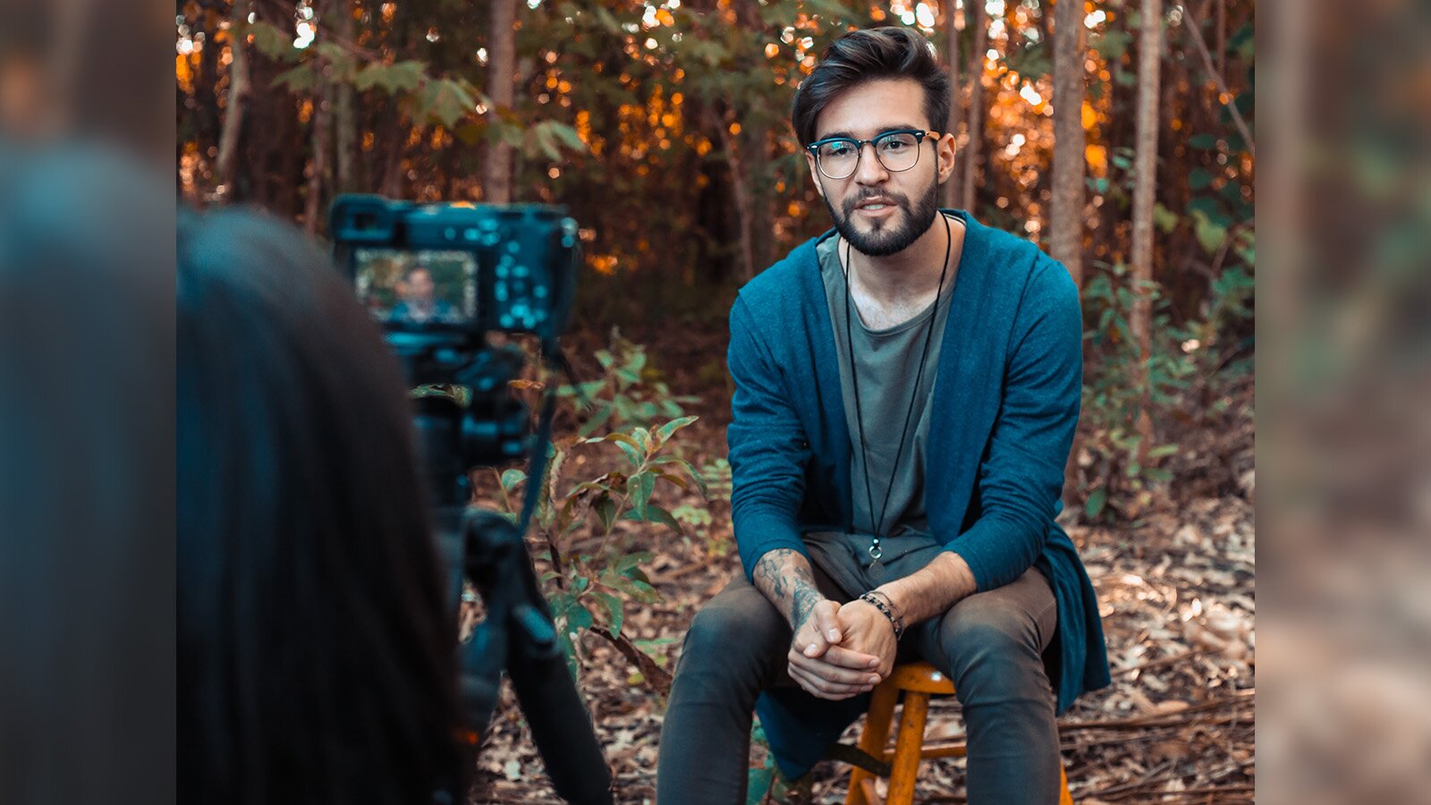 Zdjęcie przedstawia młodego mężczyznę siedzącego na taborecie na tle lasu. Przed nim ustawiono urządzenie nagrywające na statywie. 