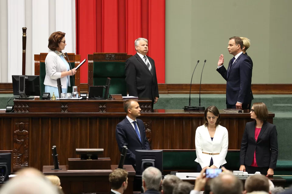 Zdjęcie przedstawia zaprzysiężenie Andrzeja Dudy na Prezydenta. Andrzej Duda wraz z małżonką stoją przy stole marszałka Sejmu. Andrzej Duda ma uniesioną prawą dłoń, powtarza za Marszałkiem Sejmu, Małgorzatą Kidawą‑Błońską, słowa przysięgi.