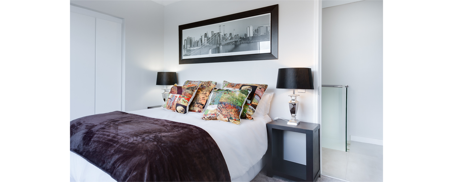 Zdjęcie przedstawia wnętrze sypialni: widoczne jest duże łóżko, po bokach lampy i stolik nocny. Nad łóżkiem wisi obraz.