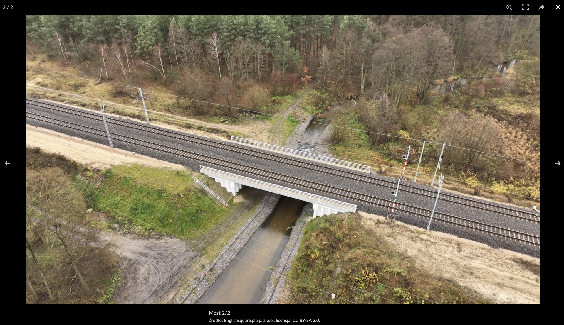 Zrzut ekranu przedstawia przykładowe zdjęcie z galerii. Wokół zdjęcia jest czarna ramka. Po prawej i lewej stronie ramki znajdują się strzałki do przewijania zdjęć w galerii. W górnym prawym rogu widoczne są cztery przyciski. Od lewej: przycisk powiększania obrazu, przycisk pełnego ekranu, przycisk udostępniania, przycisk zamknięcia podglądu zdjęcia. Pod zdjęciem widoczny jest jego podpis, a w górnym lewym rogu ramki widoczny jest numer porządkowy zdjęcia w galerii.