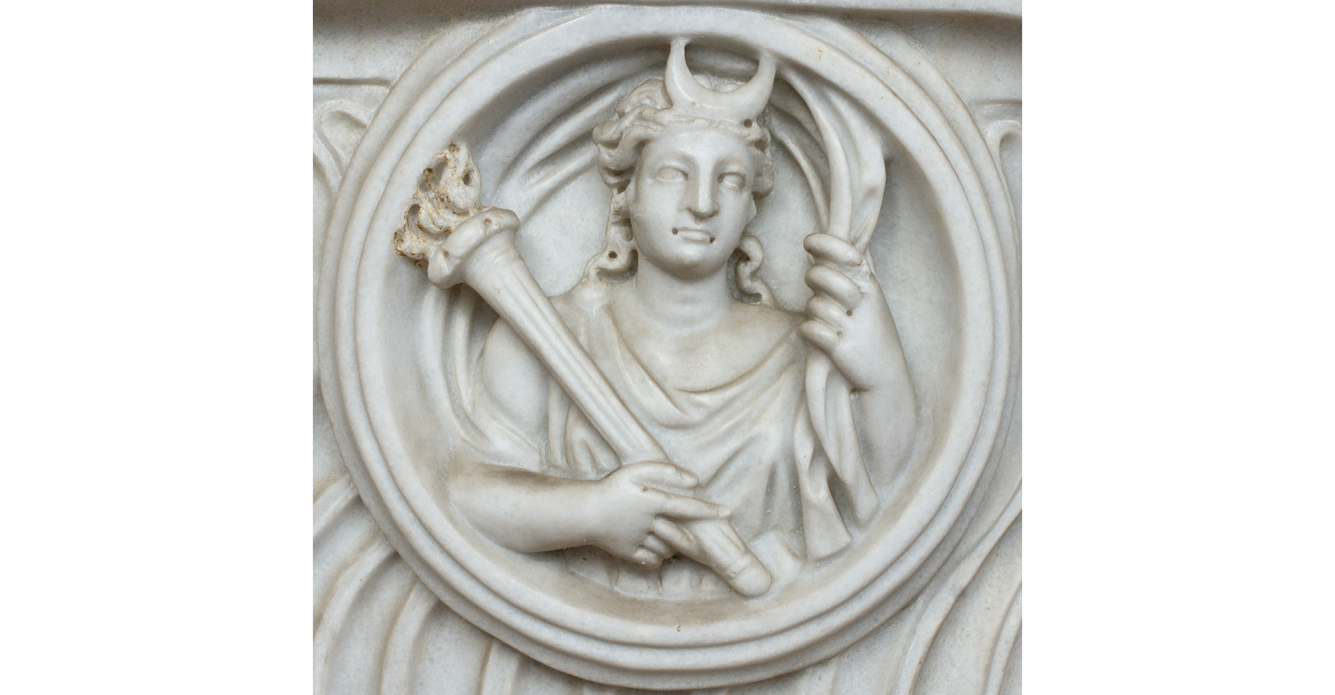 Zdjęcie przedstawia popiersie bogini księżyca Luny. Jest to marmurowa płaskorzeźba. Kobieta na głowie ma półksiężyc, a w ręku trzyma zapalony znicz.