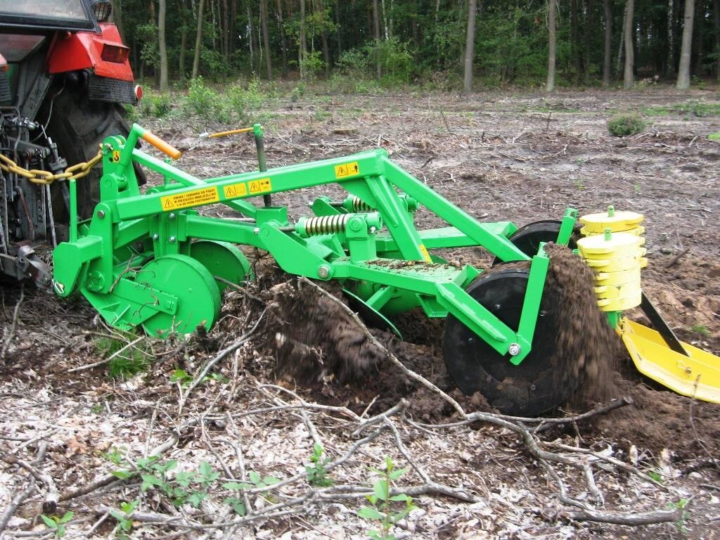 Zdjęcie przedstawia pług dwuodkładnicowy z pogłębiaczem. To metalowa rama koloru zielonego przyczepiona do tyłu ciągnika. Z obu stron ma po dwie pary kół. Z przodu, bliżej ciągnika, koła są metalowe i zielone. Po drugiej stronie koła są czarne i większe, między nimi znajdują się dwa żółte cylindry trzymające żółtą szuflę. Przekopywana ziemia wychodzi między czarnymi kołami i żółtymi cylindrami.
