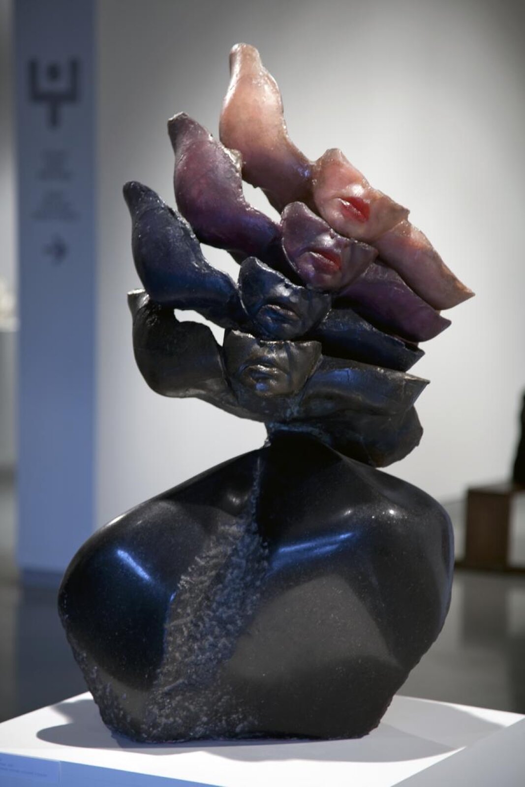 Fotografia przedstawia wieloelementową rzeźbę ułożoną na prostopadłościennej, białej podstawie na tle pomieszczenia o ciemnej posadzce i jasnych ścianach. Rzeźba to nieregularna, czarna oraz gładka bryła o kształcie niepełnego popiersia, na którym umiejscowione zostały cztery kobiecie podbródki z fragmentem pełnych ust, policzków i nosa. Na popiersiu umieszczono cztery podbródki, od dołu: czarny, ciemno fioletowy, purpurowy, lekko różowy w odcieniach skóry.