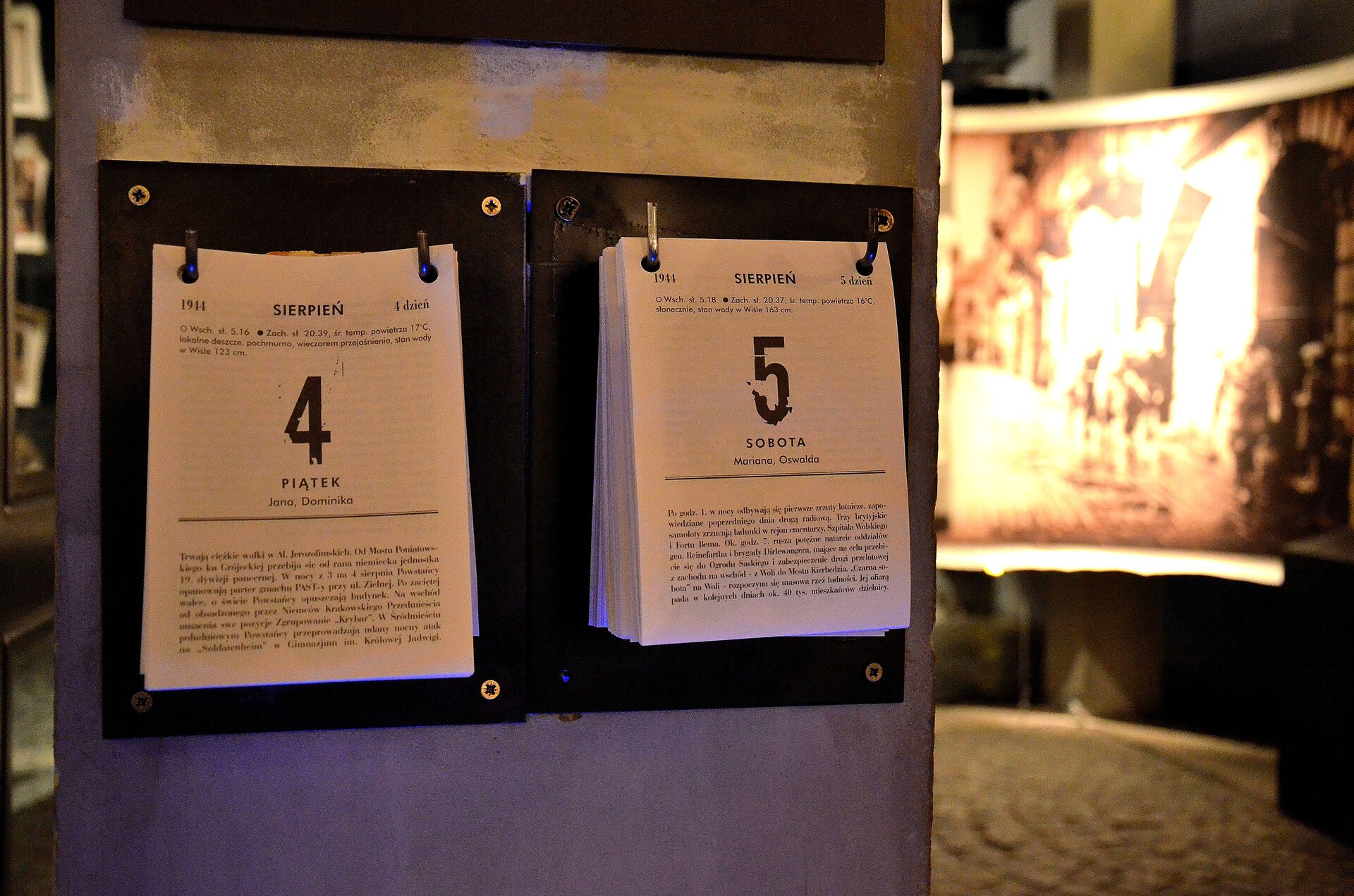 Kliknij, aby powiększyć Zdjęcie przedstawia ścianę we wnętrzu pomieszczenia. Na ścianie są zawieszone dwa grube kalendarze. Na jednym jest data 4 sierpnia 1944 roku. Na drugim 5 sierpnia 1945 roku. Pod dwoma datami, poniżej widnieją opisy walk, które miały miejsce wtedy w Warszawie.