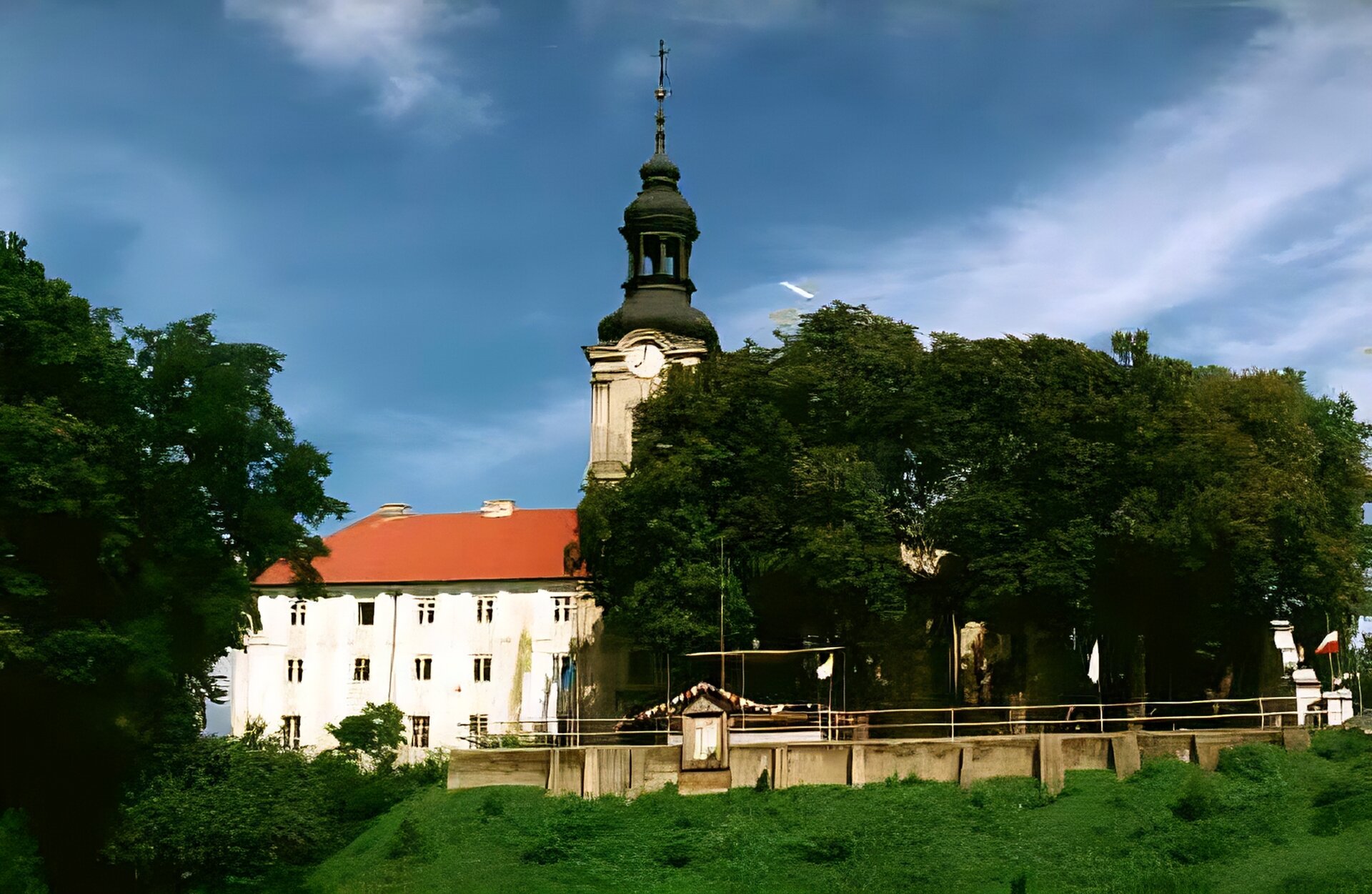 Zdjęcie przedstawia biały budynek częściowo przysłonięty przez korony drzew. Na pierwszym planie znajduje się wiele zielonych roślin oraz fragment muru. Budynek ma jasną elewacje i czerwony dach. W jego centralnej części znajduje się wieża, na szczycie której widoczny jest okrągły zegar, a nad nim szpikulec z krzyżem.