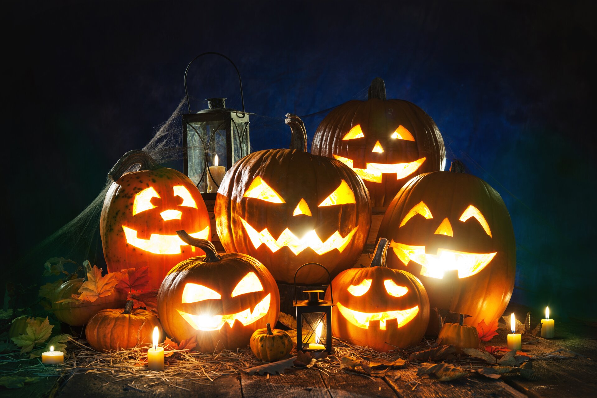 Ilustracja przedstawia sześć współcześnie wydrążonych dyń na Halloween. Są one ułożone na drewnianej podłodze. W dyniach znajdują się odpalone świece. Dookoła ustawione są lampy oraz palące się świeczki.
