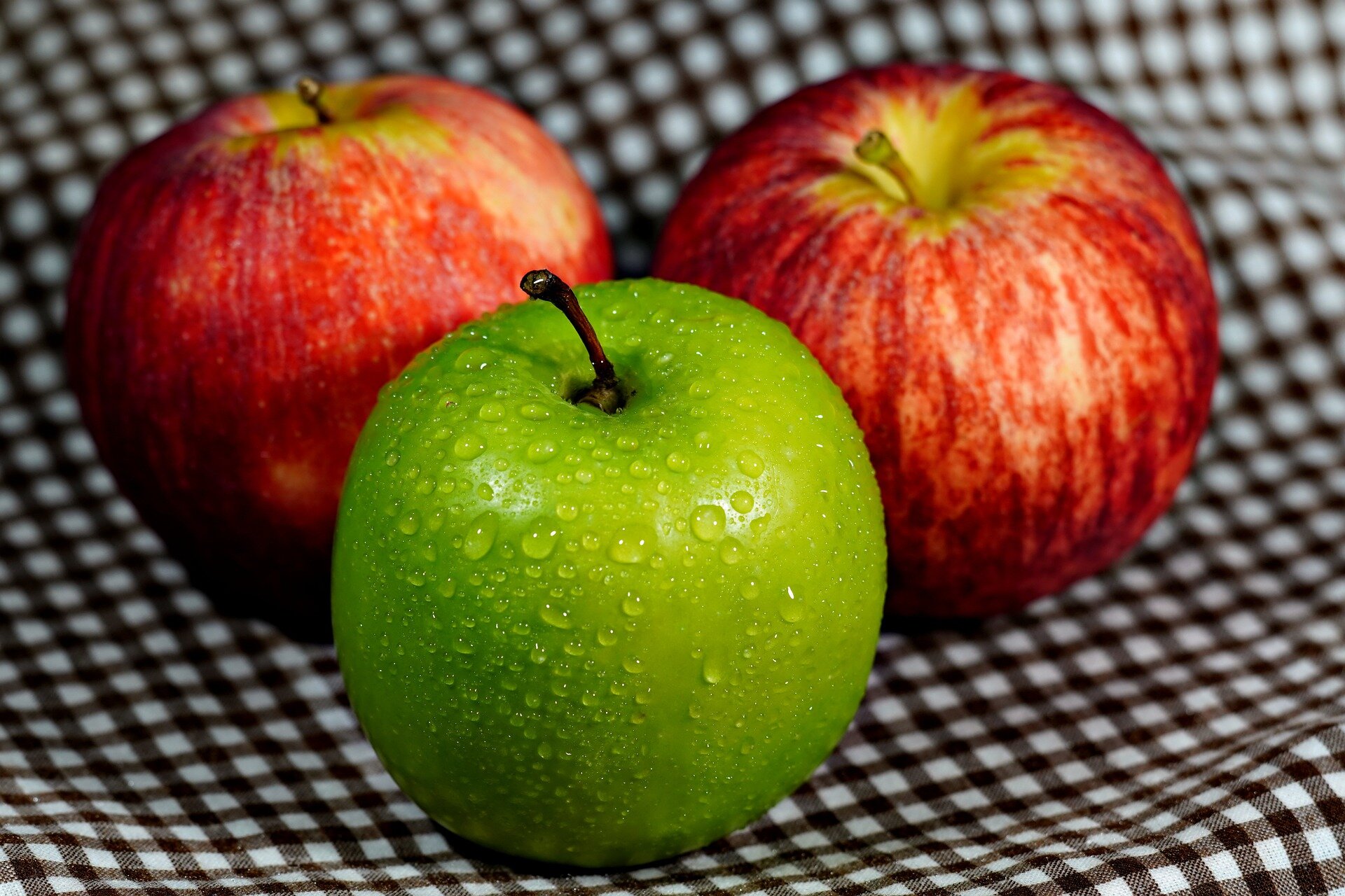 Zdjęcie przedstawia zbliżenie na leżące na obrusie trzy jabłka: dwa czerwone i jedno zielone. 
