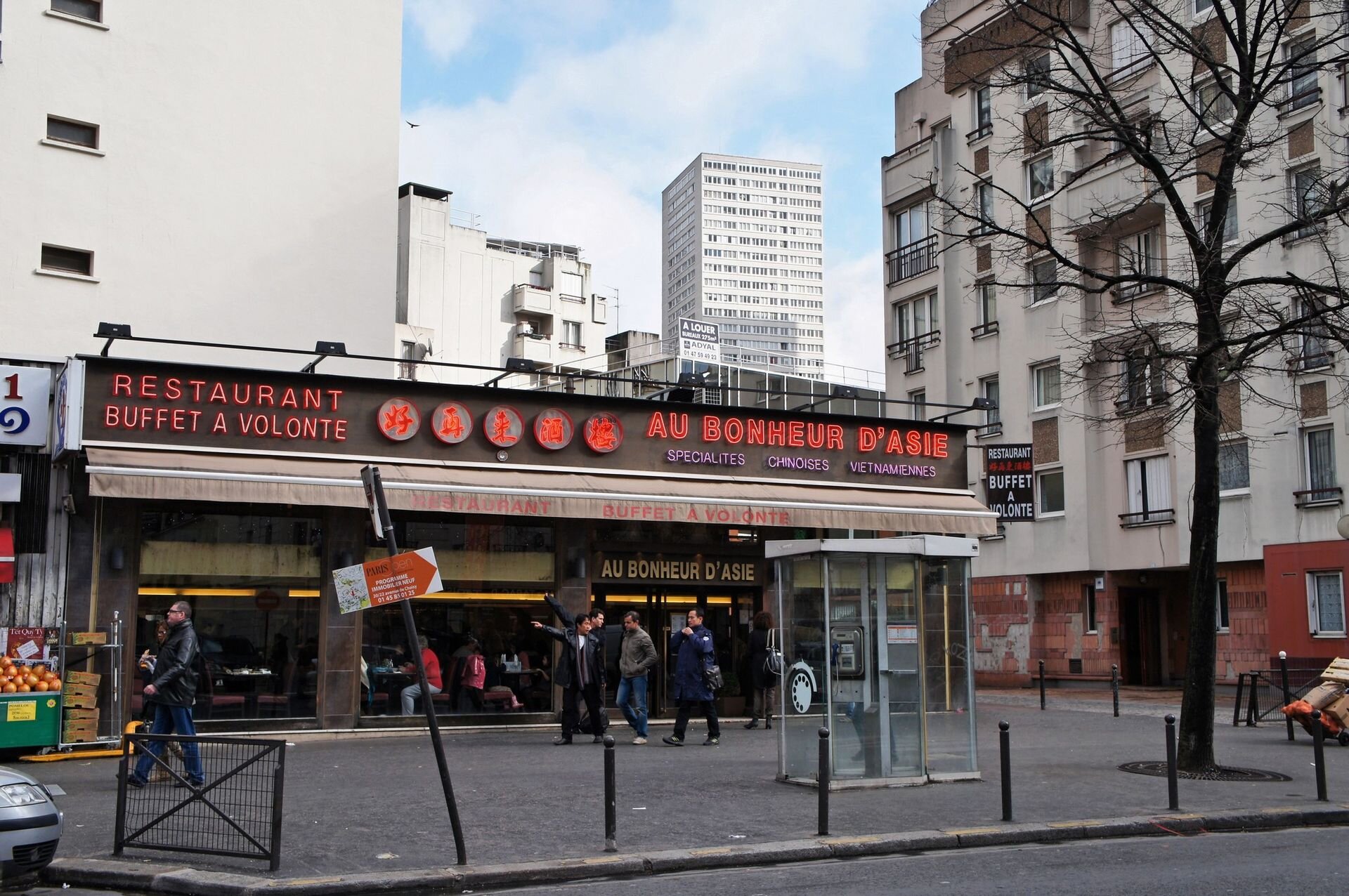 Fotografia niewielkiej restauracji położonej na rogu ulic, dookoła bloki mieszkalne, spacerowicze.