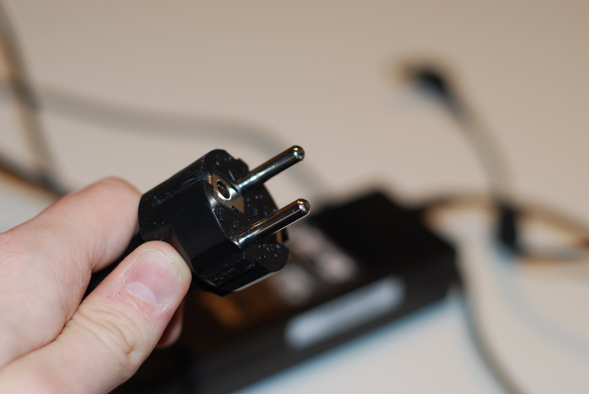Zdjęcie przedstawia standardową, europejską wtyczkę elektryczną. Wtyczka jest czarna, posiada dwa bolce, miejsce na jeden bolec z gniazdka. Jest podtrzymywana dłonią.