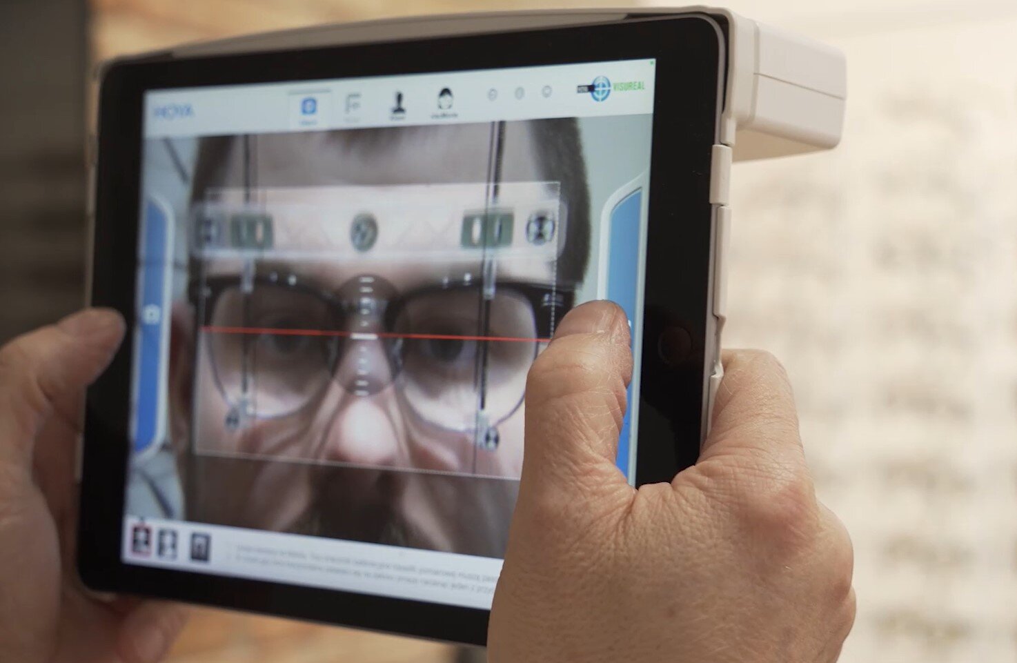 Ilustracja przedstawia wideocentrator. Jest to ekran z kamerą, na którym widać twarz badanego pacjenta. Na ekranie znajduję się czerwona linia na wysokości oczów która pomaga przy diagnozie wady.