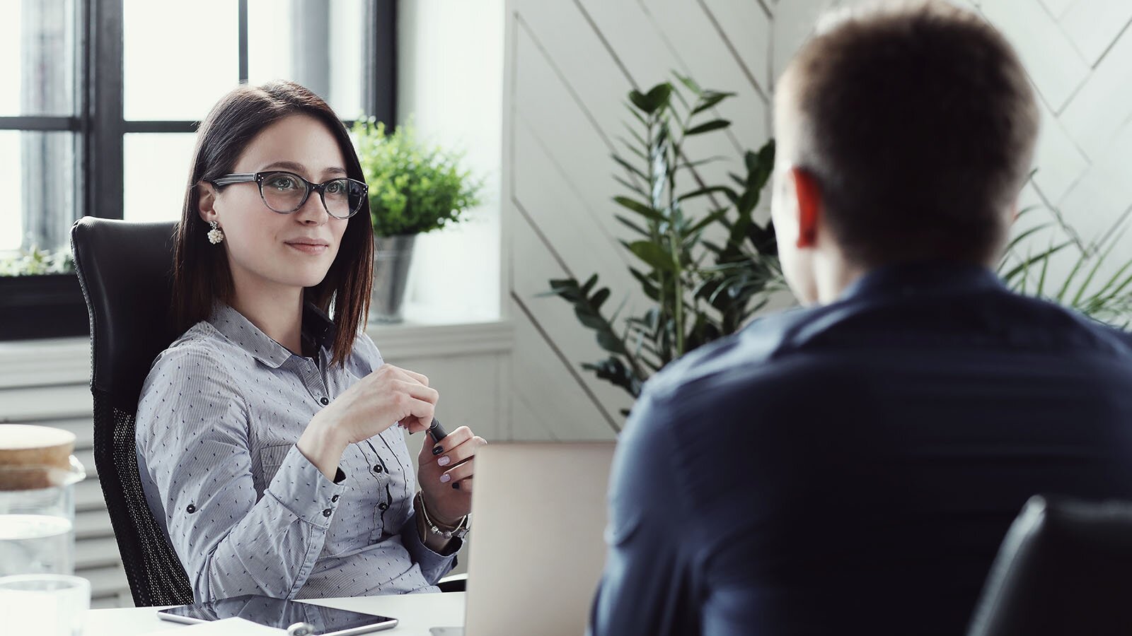 Zdjęcie przedstawia kobietę w młodym wieku siedzącą przy biurku. Przed nią po drugiej stronie biurka widoczna osoba siedząca na krześle. 