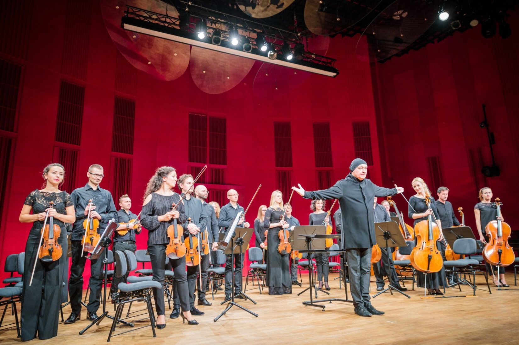 Orkiestra Kameralna Miasta Tychy pod dyrekcją Marka Mosia Fotografia przedstawia Orkiestrę Kameralną Miasta Tychy pod dyrekcją Marka Mosia na scenie. Parkiet jest jasnobrązowy, a ściana sali koncertowej została podświetlona na czerwono. Z przodu stoi mężczyzna ubrany na czarno, który wyciąga ręce na boki. Za nim jest około dwudziestu muzyków, z których większa część trzyma przed sobą w dłoniach brązowe skrzypce i smyczki. Z prawej strony stoją muzycy trzymający brązowe wiolonczele. Muzycy ubrani są w czarne stroje, większość z nich patrzy przed siebie. Obok nich stoją ciemne krzesła oraz pulpity na nuty.