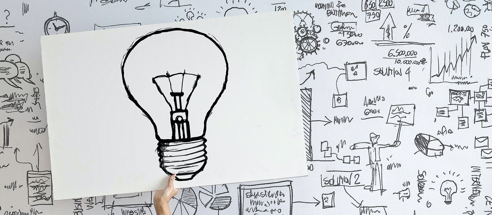 Grafika przedstawia trzymany w dłoni odręczny rysunek żarówki, który przedstawiony jest na powierzchni zapełnionej odręcznymi rysunkami, dymkami, schematami, wzorami i obliczeniami. 