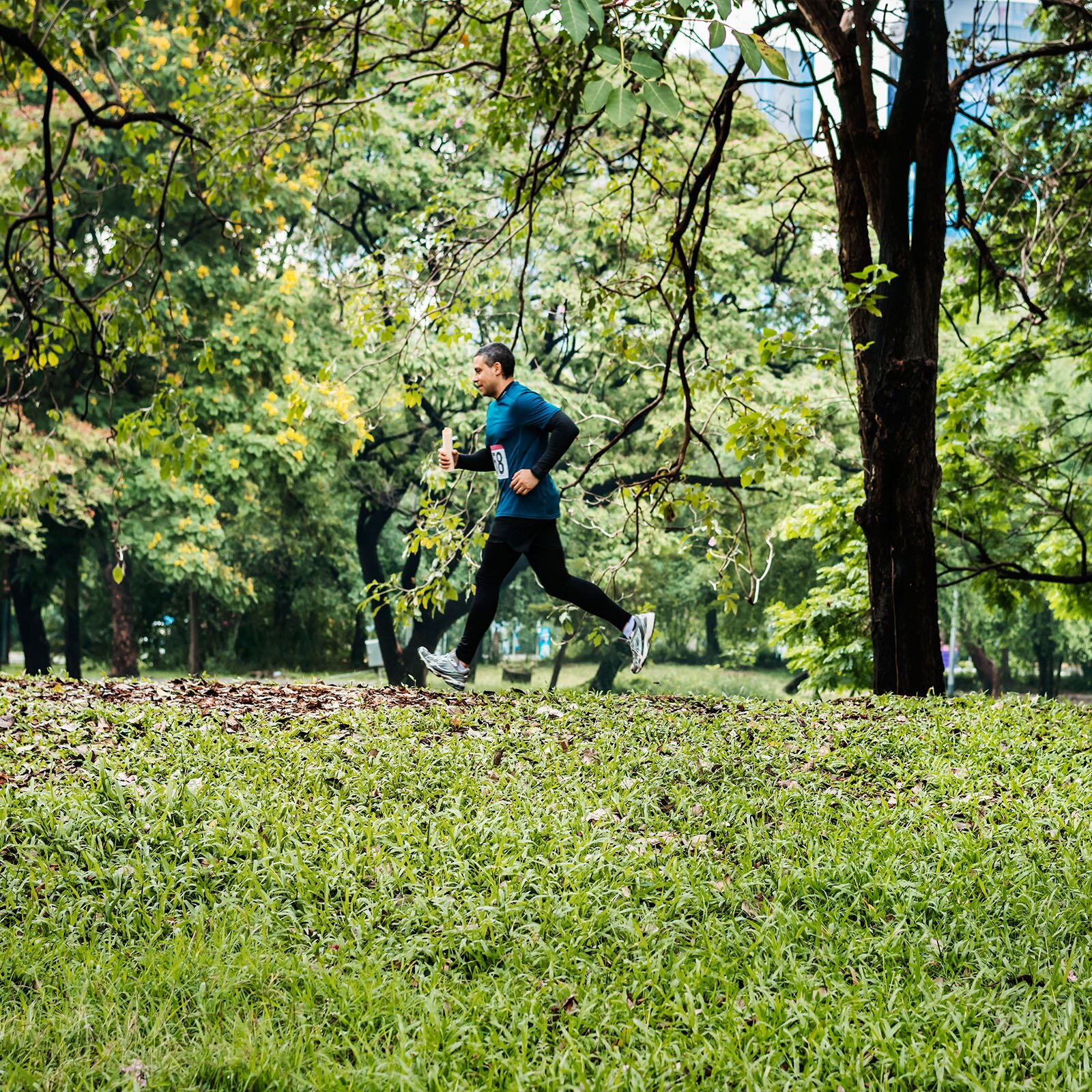  Zdjęcie przedstawia osobę biegnącą przez park, otoczoną zielenią. W tle widoczne są drzewa, liście oraz fragmenty miejskiego krajobrazu. Biegacz ma na sobie niebieską koszulkę i czarne legginsy, a na stopach sportowe buty do biegania. Wydaje się być zaangażowany w aktywność sportową na świeżym powietrzu.