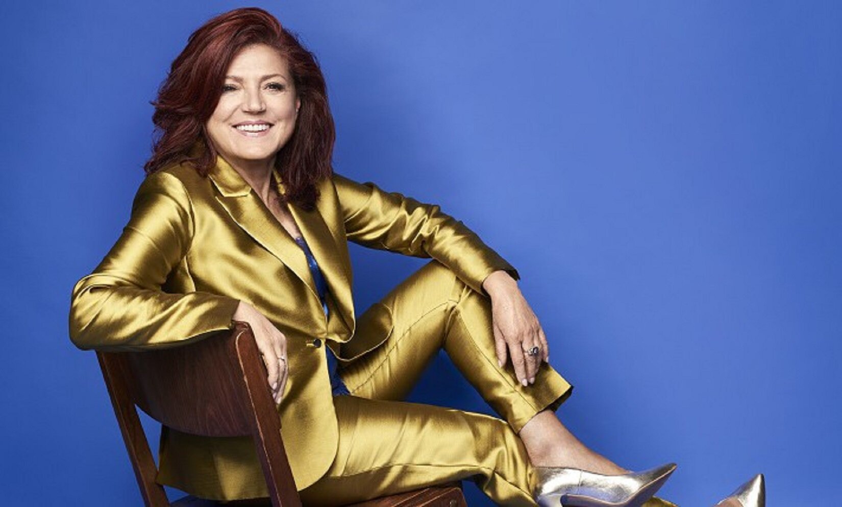 Fotografia przedstawia uśmiechniętą kobietę siedzącą na krześle. Kobieta jest w starszym wieku, ma na sobie błyszczący złoty garnitur, błyszczące srebrne szpilki oraz ciemnoniebieską bluzkę, ledwo widoczną spod zapiętej na guzik marynarki. Kobieta siedzi na ciemnobrązowym drewnianym krześle, opiera się na nim prawym łokciem i lekko wychyla się z krzesłem do tyłu. Na fotografii przedstawiona jest bokiem do aparatu. Lewą nogę ma założoną na prawą. Uśmiecha się szeroko. Na jej twarzy malują się zmarszczki. Ma bujne, brązowe włosy. Tło jest niebieskie.