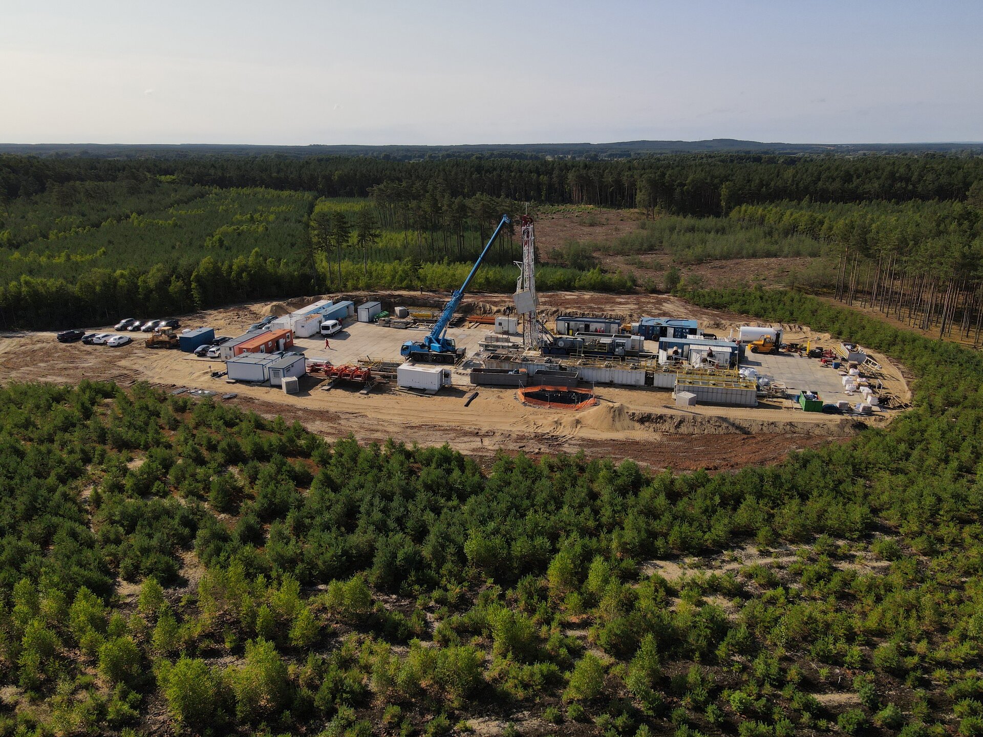 Na zdjęciu widać maszt wiertniczy znajdujący się na placu wiertniczym widoczny z góry.Plac wiertniczy widoczny jest z lotu ptaka. Ma kształt prostokąta. Podstawa placu jest wyłożona betonowymi płytami. W centralnym punkcie placu znajduje się maszt wiertniczy. Maszt jest w kształcie ostrosłupa ściętego. Maszt znajduje się pomiędzy białymi kontenerami. Po lewej stronie masztu widoczny jest niebieski dźwig. Przed placem wiertniczym znajduje się parking samochodowy.W tle zdjęcia widać tereny leśne.