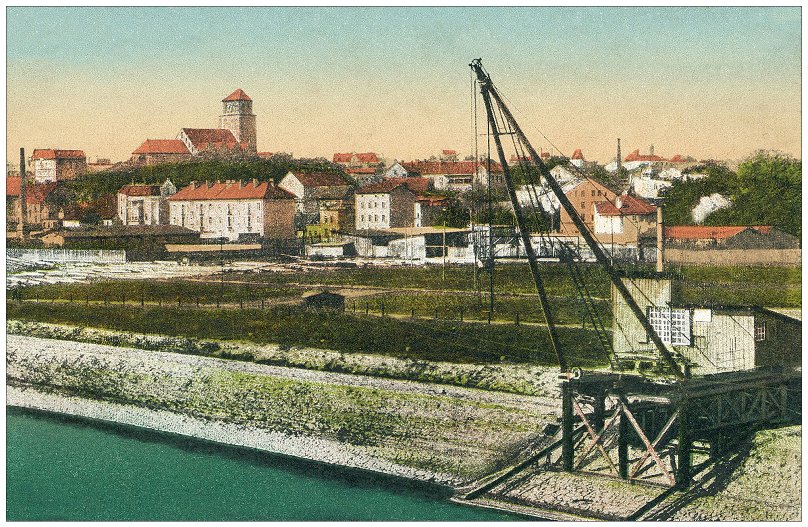 Zdjęcie przedstawia widok na Tczew od strony Lisewa. Na pierwszym planie, rzeka z nabrzeżem, po prawej stronie zdjęcia jest usytuowany fragment portu rzecznego z rampą przeładunkową i dźwigiem. Dalej zielone pola. Na drugim planie budynki, kilkupiętrowe, murowane, ze skośnymi dachami i kominami. Po lewej stronie niższy, murowany budynek oraz drugi z wysokim kominem. Po lewej stronie widoczny jest gotycki kościół z wysoką, szeroką wieżą.