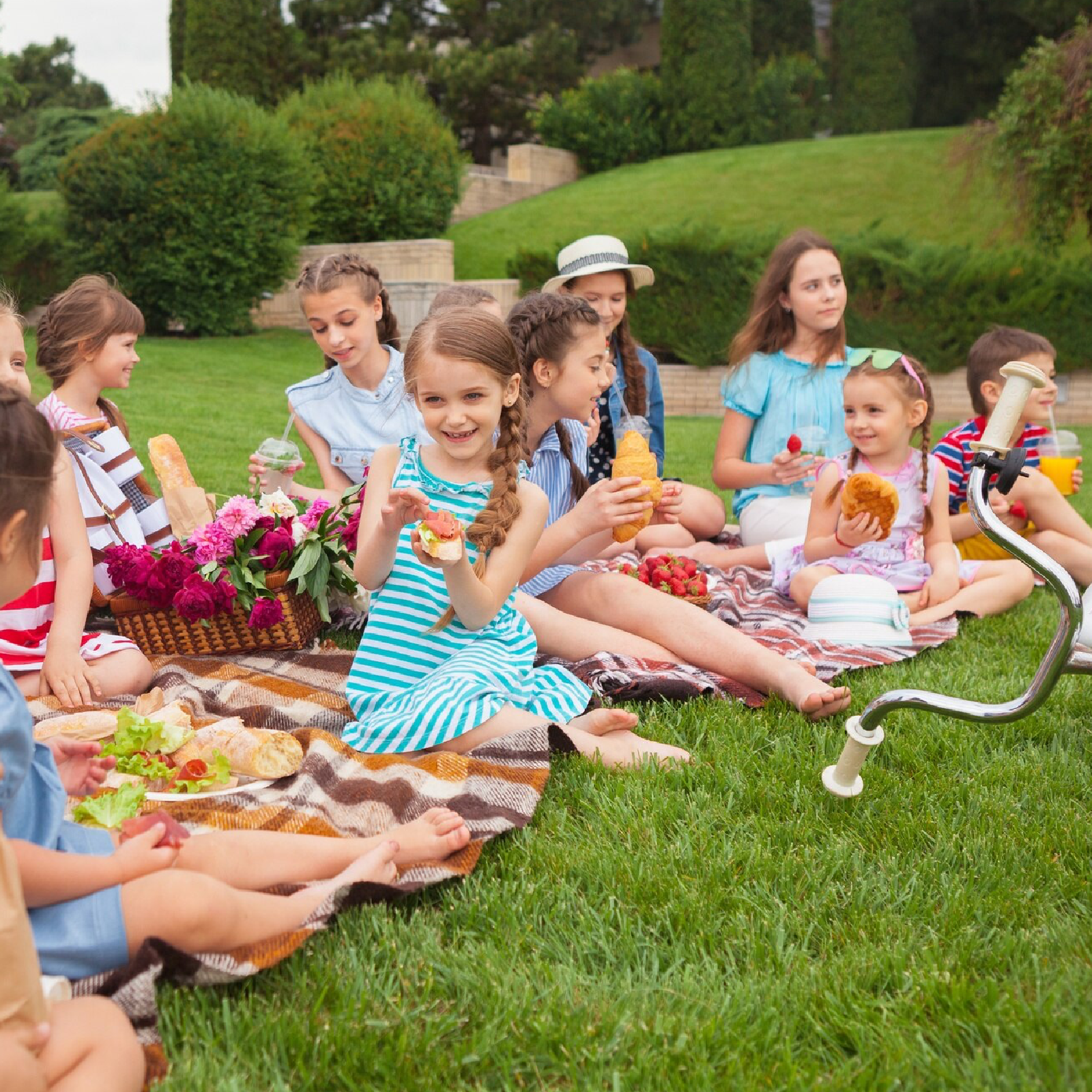 Zdjęcie przedstawia dzieci w wieku szkolnyym na pikniku, na trawie. Siedzą na kocach i jedzą. 