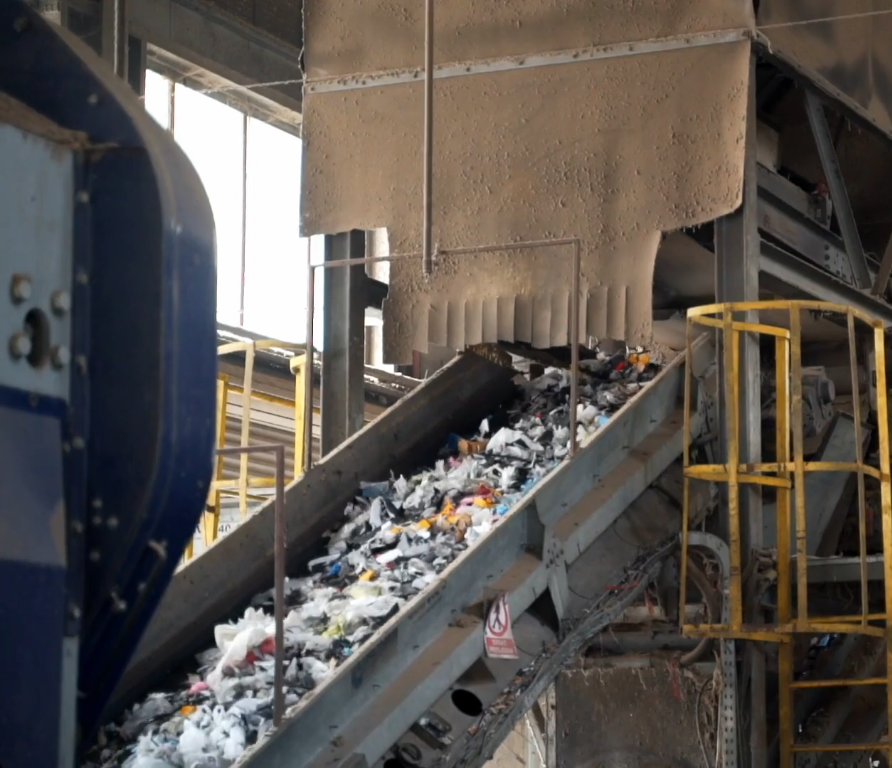 Zdjęcie przedstawia taśmociąg kanałowy. Znajduje się on w hali i ustawiony jest pod kątem ok 25 stopni. Składa się z przesuwnej taśmy z wysokimi bocznymi ściankami, którą odpady przemieszczają się z miejsca na miejsce w obrębie hali.