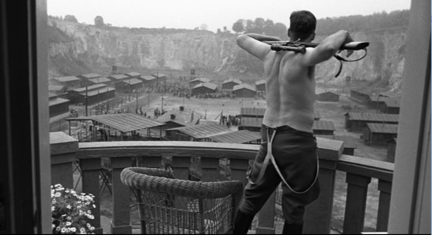 Zdjęcie przedstawia Gӧtha stojącego na balkonie, w samych wojskowych spodniach. Mężczyzna trzyma na karku karabin. Patrzy się na plac obozowy pełen baraków.