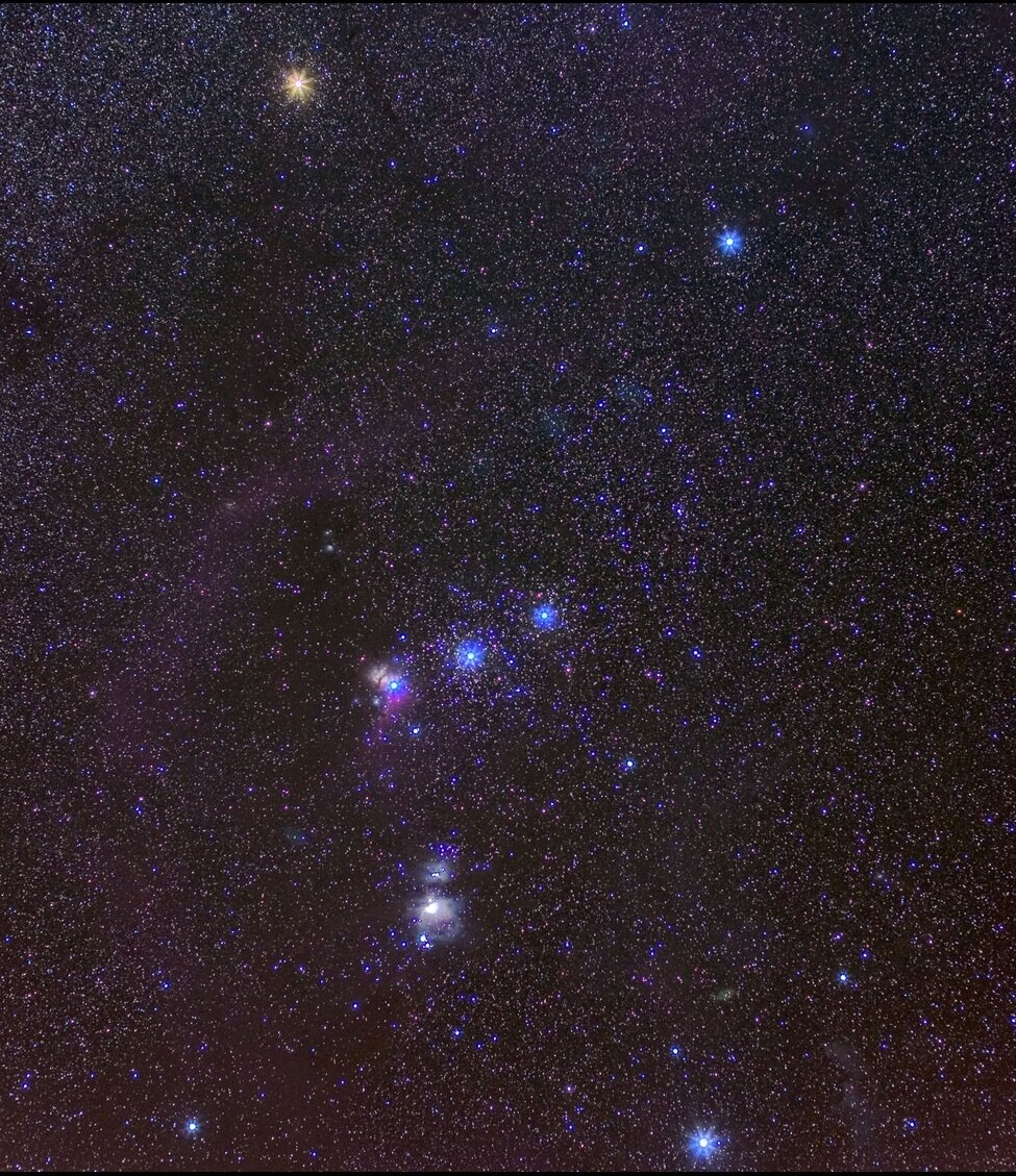 Fotografia prezentuje gwiazdozbiór Oriona. Na granatowym niebie jest kilka dużych, jasnych łatwo rozpoznawalnych gwiazd. Pas Oriona przypomina kształtem pochyloną w prawo dużą literę T.