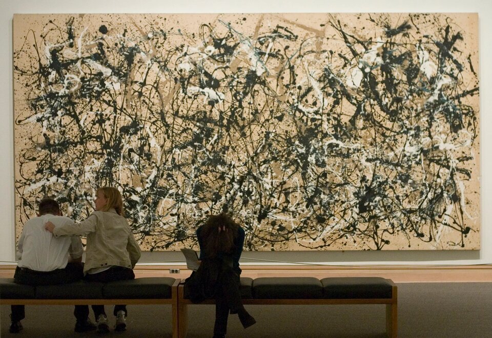 Fotografia przedstawia wielki obraz oraz trzy znajdujące się przed nim osoby. Obraz wykonany jest w stylu abstrakcyjnym, ma układ poziomy i składa się z plątaniny wielu nieregularnych linii różnej grubości, nachodzących na siebie. Obraz jest dynamiczny, bardzo dużo się na nim dzieje, a linie są w kolorze czarnym, białym i brązowym na jasnobrązowym tle.  Pomieszczenie nie jest zbyt jasno oświetlone. Dzieło znajdujące się na ścianie ma układ poziomy i wykonane jest w stylu abstrakcyjnym. Przed obrazem stoją dwie drewniane ławki z ciemnymi, tapicerowanymi siedzeniami. Na ławce znajdującej się po lewej stronie, przodem do obrazu, siedzą dwie osoby - kobieta i mężczyzna. Mężczyzna ma ciemne włosy, ubrany jest w białą bluzę i ciemne spodnie, nogi ma rozstawione szeroko, a łokcie opiera na kolanach. Siedząca obok niego kobieta ubrana jest w beżową kurtkę, granatowe spodnie oraz jasne, sportowe buty. Kobieta trzyma swoją lewą rękę na plecach mężczyzny, a wzrok skierowany ma w lewo. Na drugiej ławce siedzi kobieta skierowana plecami do obrazu. Kobieta ma na sobie ciemne ubrania, nogę założoną na drugą nogę oraz skrzyżowane ręce oparte na kolanach. Głowę pochyla nisko, w jednej ręce trzyma kartkę papieru.