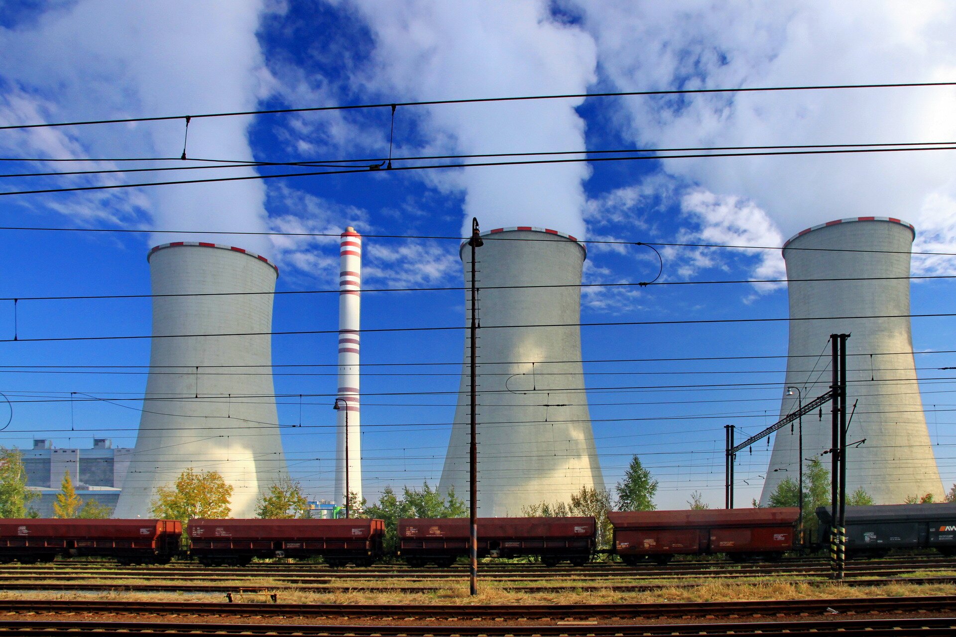 Fotografia prezentuje fragment elektrowni na węgiel kamienny. Widoczne trzy białe duże kominy, z których wydobywa się para wodna. Przed nimi tory kolejowe, którymi przejeżdża pociąg.