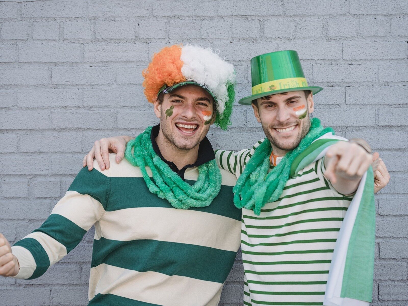 Zdjęcie przedstawia dwóch mężczyzn na tle ściany z białej cegły ubranych w barwy Irlandii (zielony, biały, pomarańczowy). Jeden mężczyzna ma na głowie kapelusz a drugi perukę, obaj mężczyźni są uśmiechnięci.