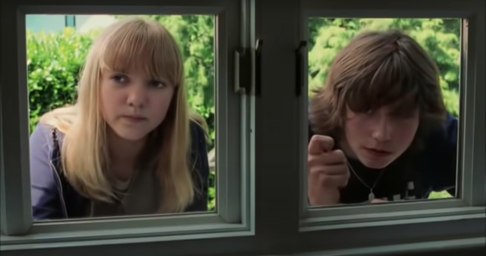 Zdjęcie przedstawia okno, za którym, na dworze stoi dziewczynka i chłopiec. W tle zielone krzewy.