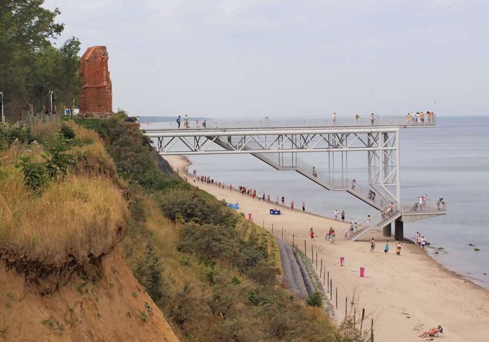 Zdjęcie pokazuje ruiny kościoła w Trzęsaczu z perspektywy bocznej. Na brzegu urwiska po lewej stronie zdjęcia bokiem do patrzącego stoi czerwona, ceglana ściana. Poniżej zbocze urwiska z porastającymi je roślinami, które na dole łączy się z nadmorską plażą. Na poziomie ruin kościoła taras widokowy wysunięty w morze, wsparty na metalowych, białych palach. Z plaży na taras prowadzą schody. Na tarasie i na plaży znajdują się ludzie. Po lewej stronie morze. 