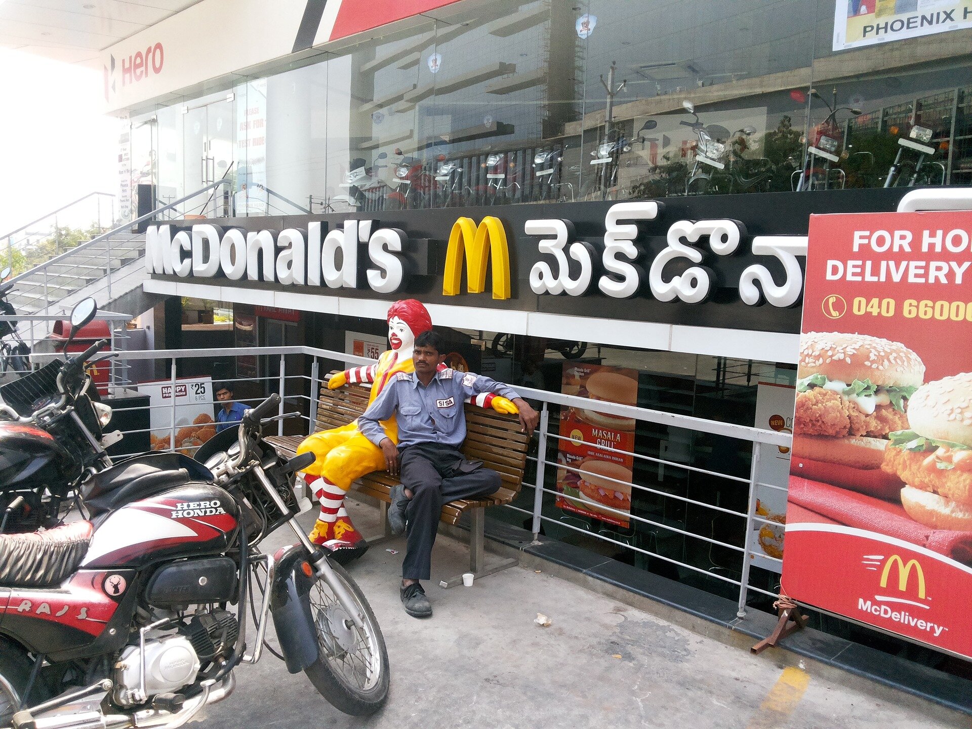 Na zdjęciu restauracja McDonalds w Indiach. Przed restauracją na ławce siedzi mężczyzna o ciemnej karnacji – Hindus. Obok niego plastikowa maskotka restauracji – klaun naturalnych rozmiarów człowieka. Przed restauracją zaparkowane motocykle.