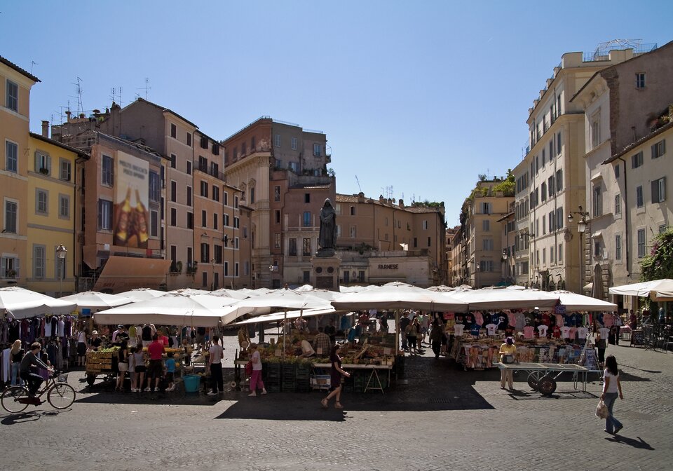 Kliknij żeby powiększyć Zdjęcie przedstawia plac Campo di Fiori w Rzymie. Na środku stoi pomnik Giordana Bruna. Wokół znajdują się kamienice. Wokół pomnika są rozstawione stragany, przy których stoją ludzie.