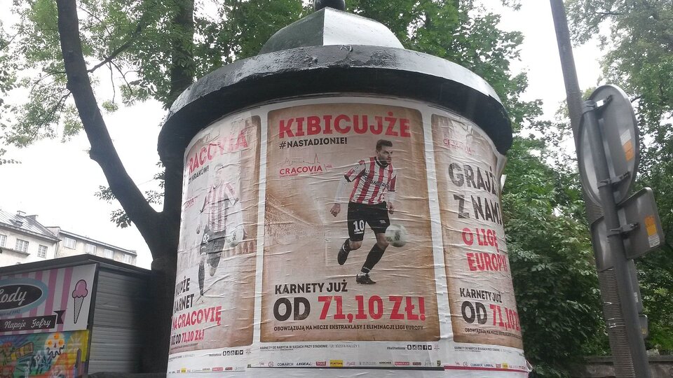 Zdjęcie przedstawia słup ogłoszeniowy, na którym przyklejony jest plakat. Na plakacie widzimy piłkarza w stroju sportowym oraz z piłką przy nodze. Na górze plakatu napis „kibicujże”. Na dole napis „Karnety już od 71,10 zł”.