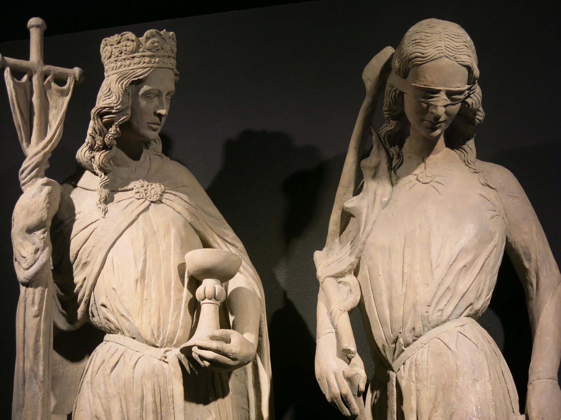 Zdjęcie przedstawia dwie rzeźbione figury kobiece. Po lewej stronie znajduje się Eklezja. To młoda kobietra w długiej szacie i pelerynie, wspierająca się na trzymanym prawą ręką krzyżu. Kobieta trzyma w lewej ręce kielich. Na głowie ma królewską koronę. Kobieta po prawej stronie także jest młoda, w długiej sukni. Ma zawiązane oczy.